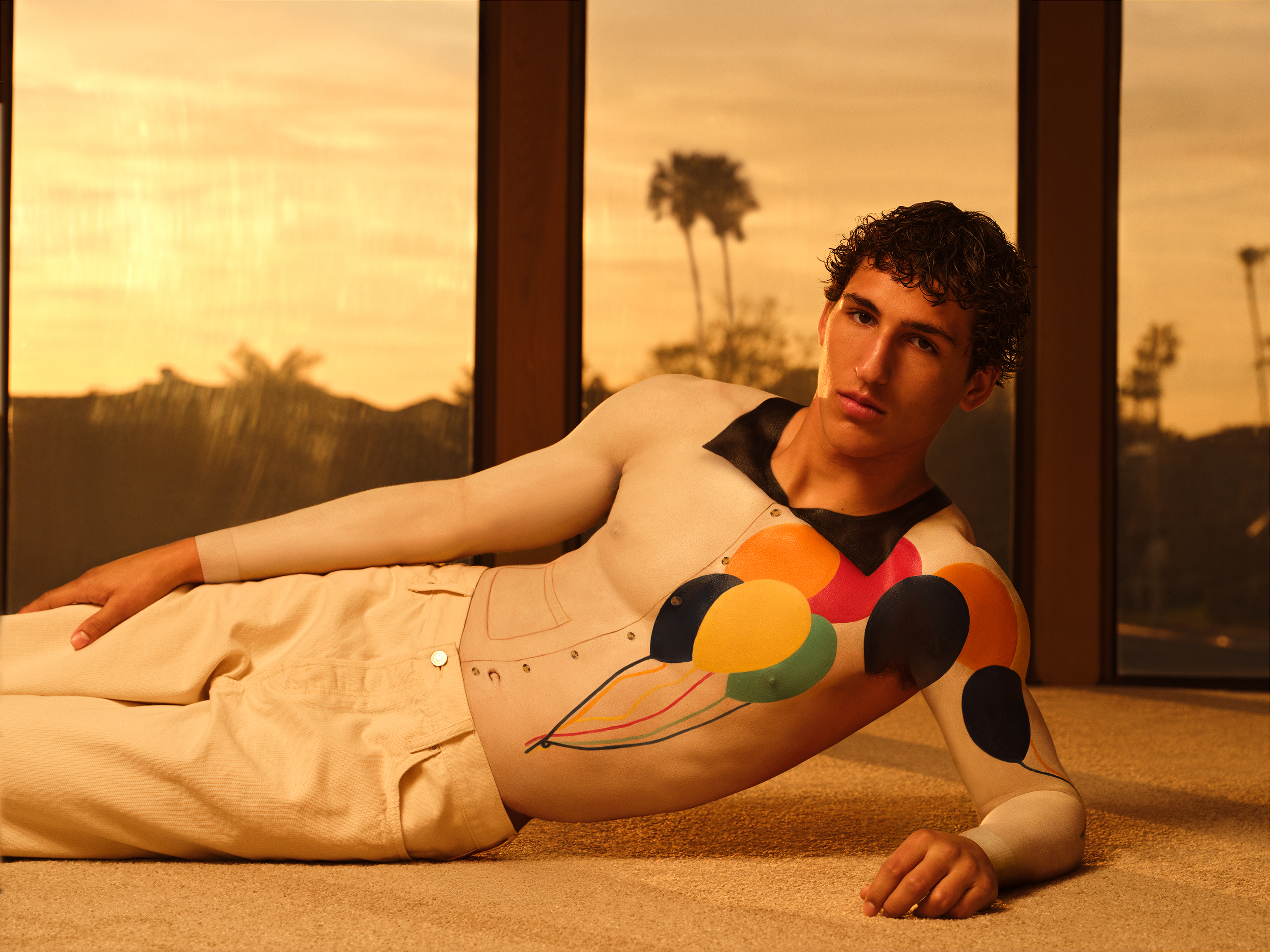 Male model reclined on carpet looking at the camera with a white body painted top with baloon details against a palm tree backdrop
